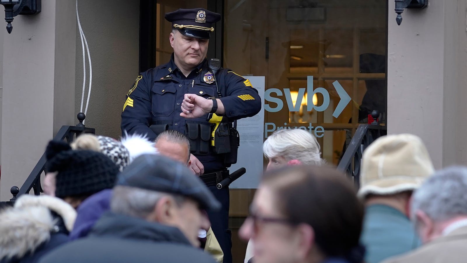 A Wellesley, Mass., police officer glances at his watch as customers and bystanders form a line outside a Silicon Valley Bank branch location, Monday, March 13, 2023, while waiting for the branch to open, in Wellesley, Mass. Photo by AP.